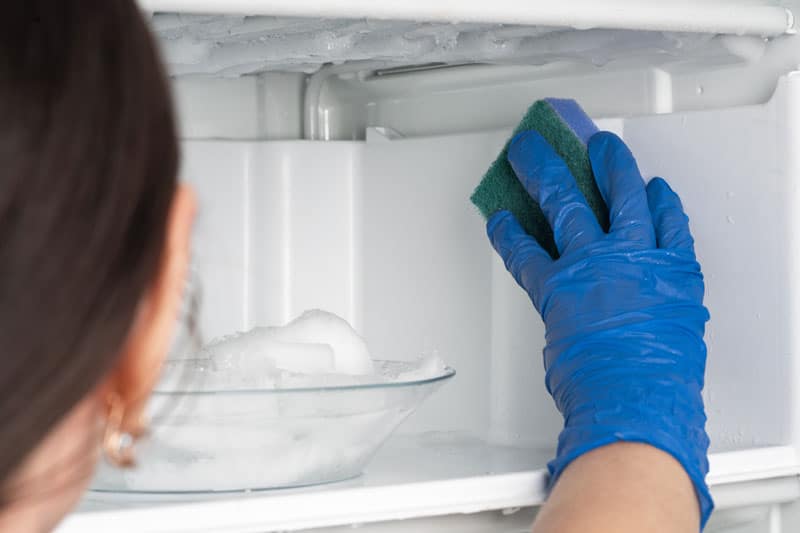 Femme qui dégivre et nettoie son frigo à l'aide d'une éponge